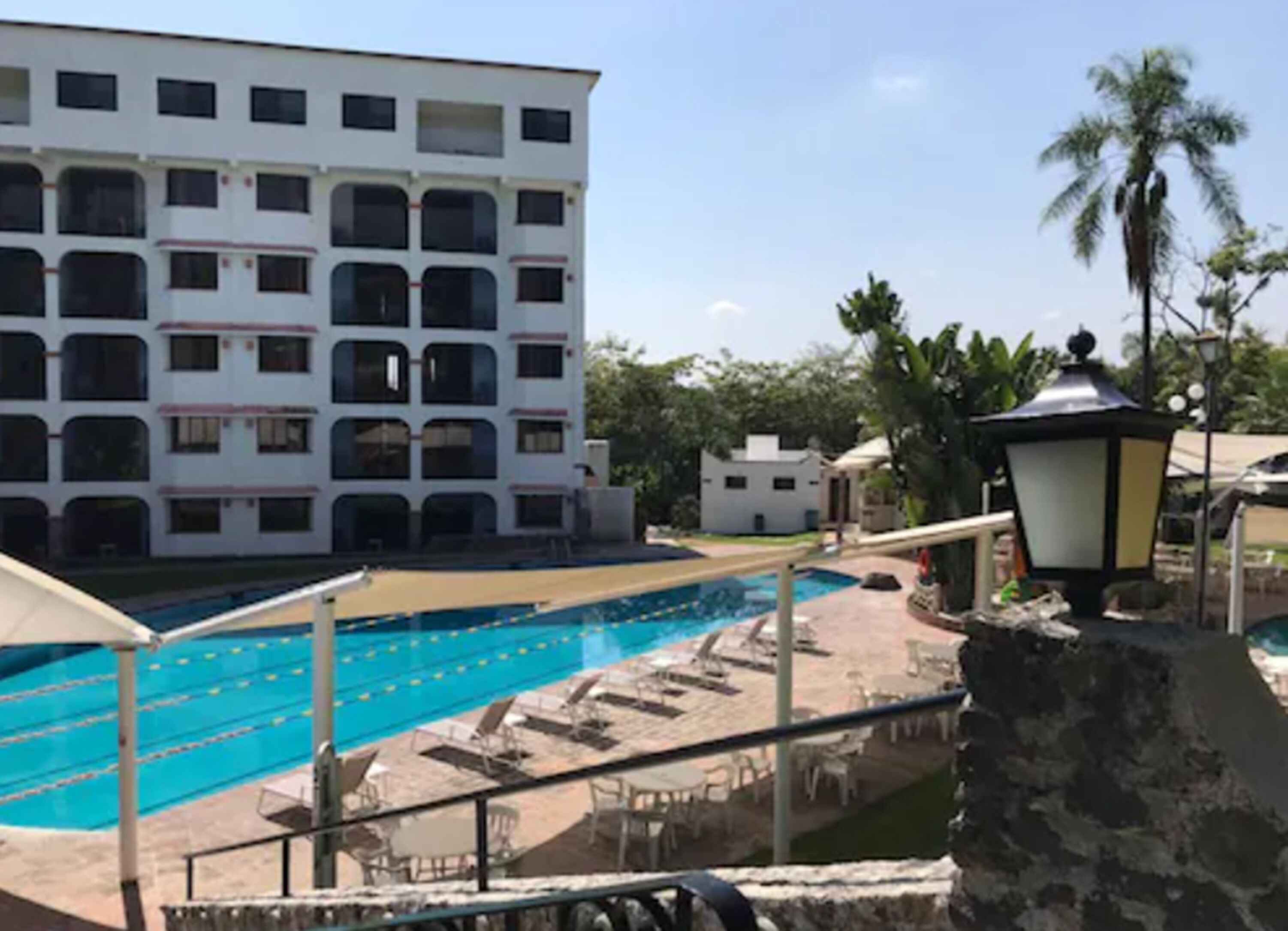 Una piscina con varias pistas, un edificio blanco con balcones y una palmera.