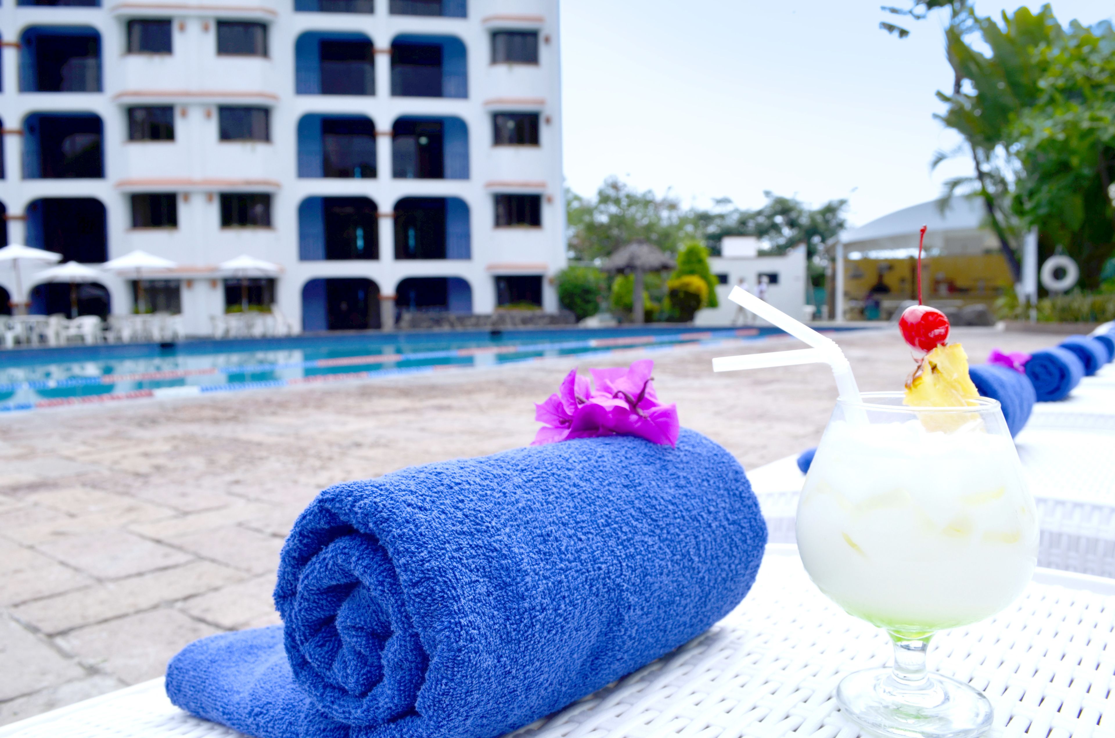 A poolside setting with a blue towel, a drink with a cherry, and a building with multiple windows in the background.