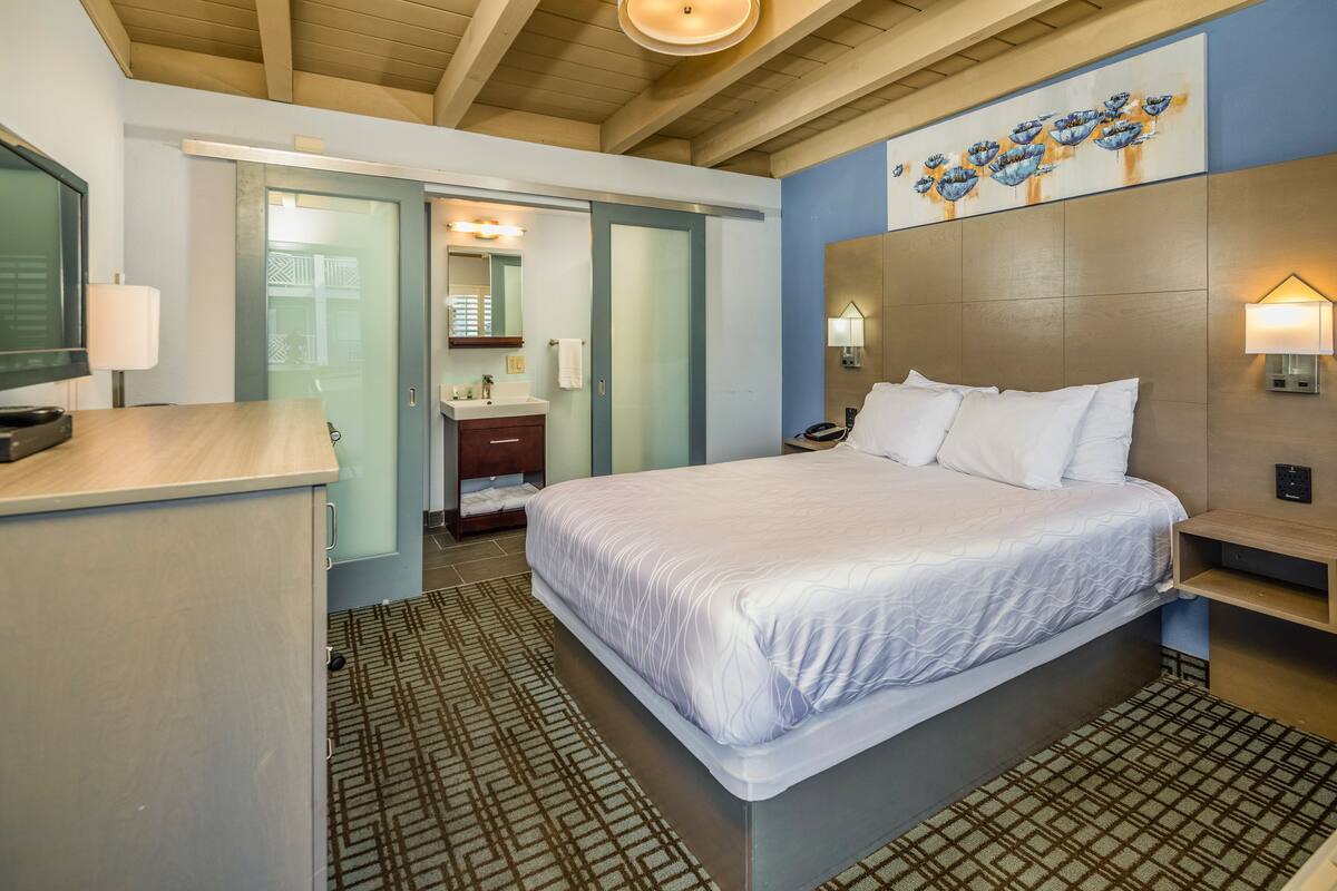A hotel room with a bed, bedside table, lamp, and a bathroom with a sink and mirror.