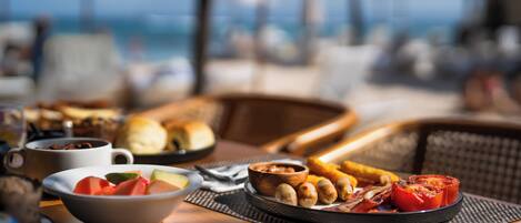 A breakfast table with various food items, including fruits, bread, and a bowl of nuts, set on a wooden table with a view of the sea in the background.