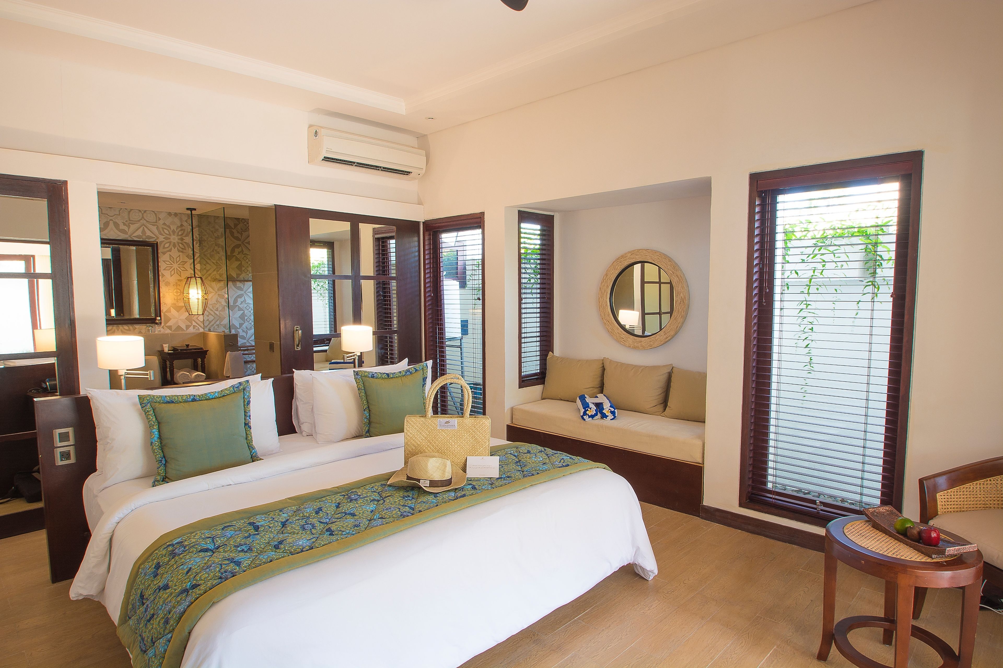 Une chambre d’hôtel avec un grand lit, un canapé, un miroir rond et une fenêtre avec des stores.