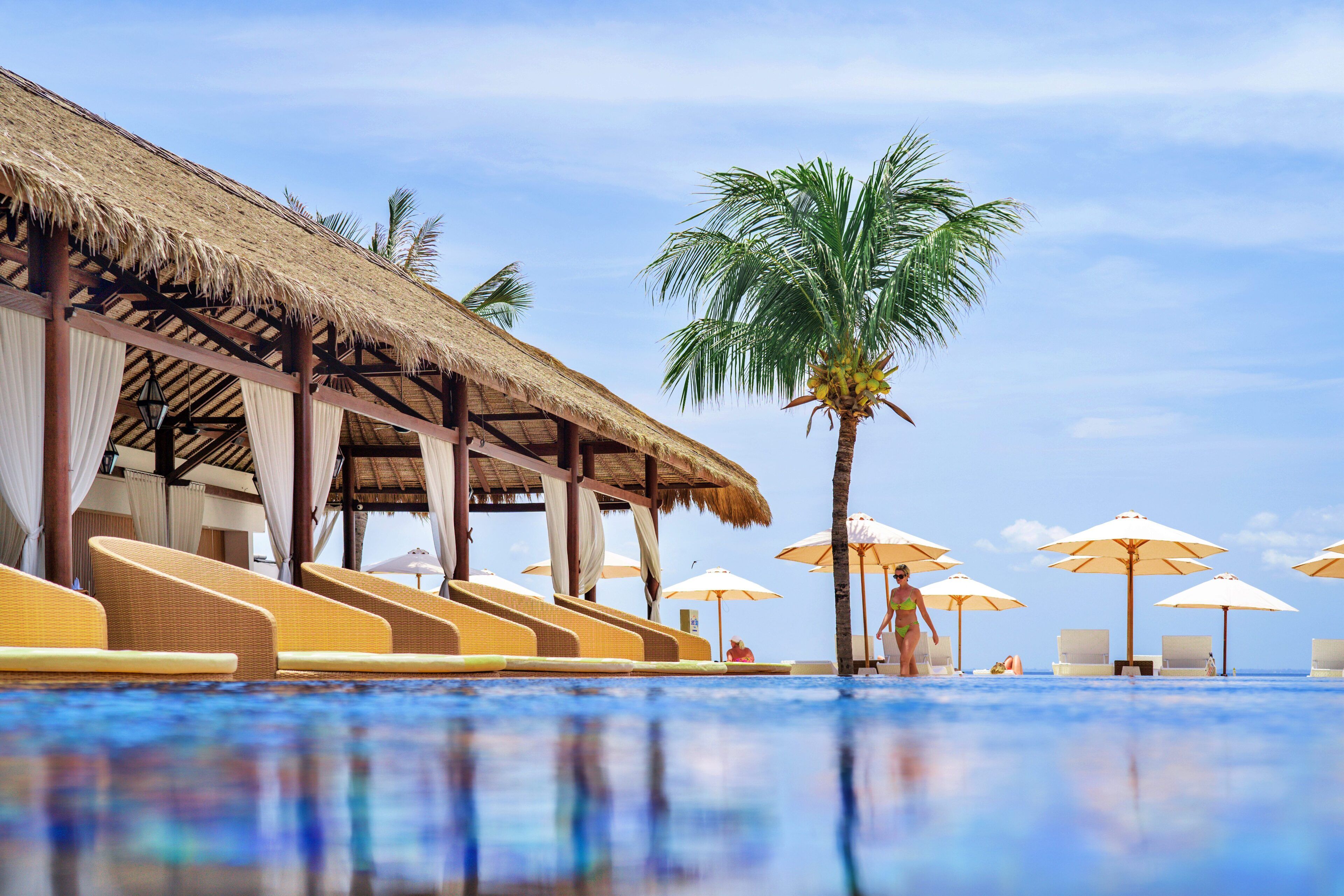 Uma área ao lado da piscina com espreguiçadeiras e guarda-sóis, uma construção com telhado de palha e palmeiras.