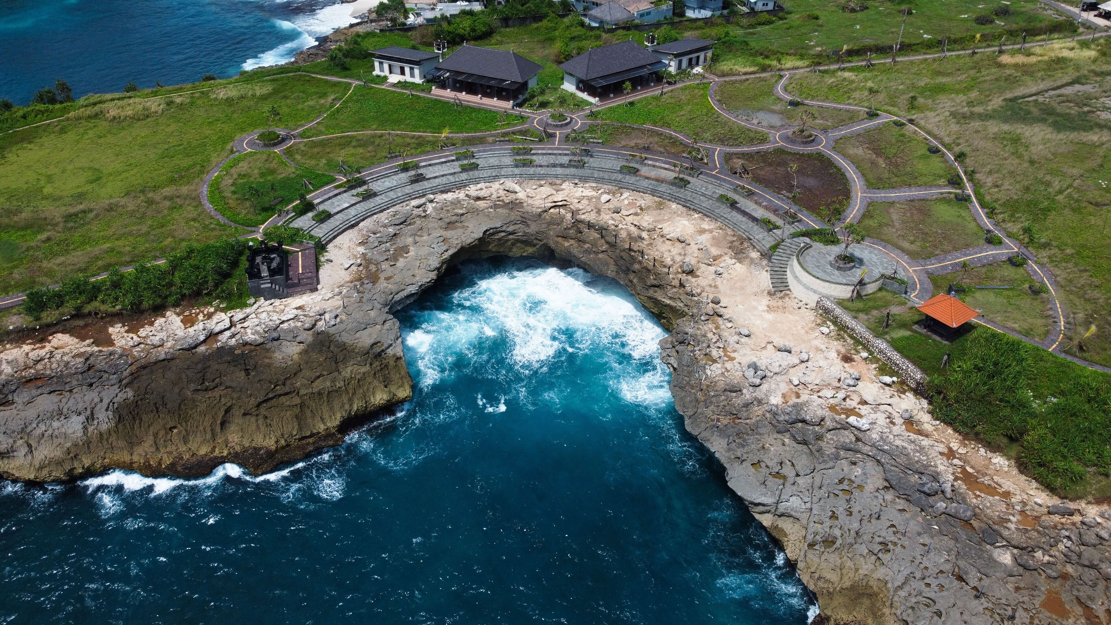 Une vue aérienne d’une zone côtière, avec une formation rocheuse imposante s’élevant en arc au-dessus de l’océan, une route sinueuse et plusieurs bâtiments aux toits distinctifs.