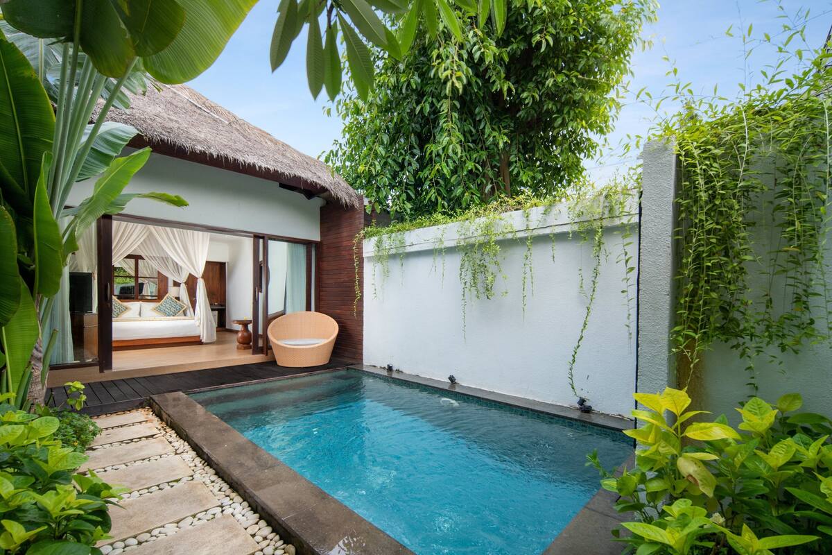 Un espace piscine agrémenté d’une structure à toit de chaume, d’un mur blanc recouvert de plantes grimpantes et d’un lit avec du linge de maison blanc.