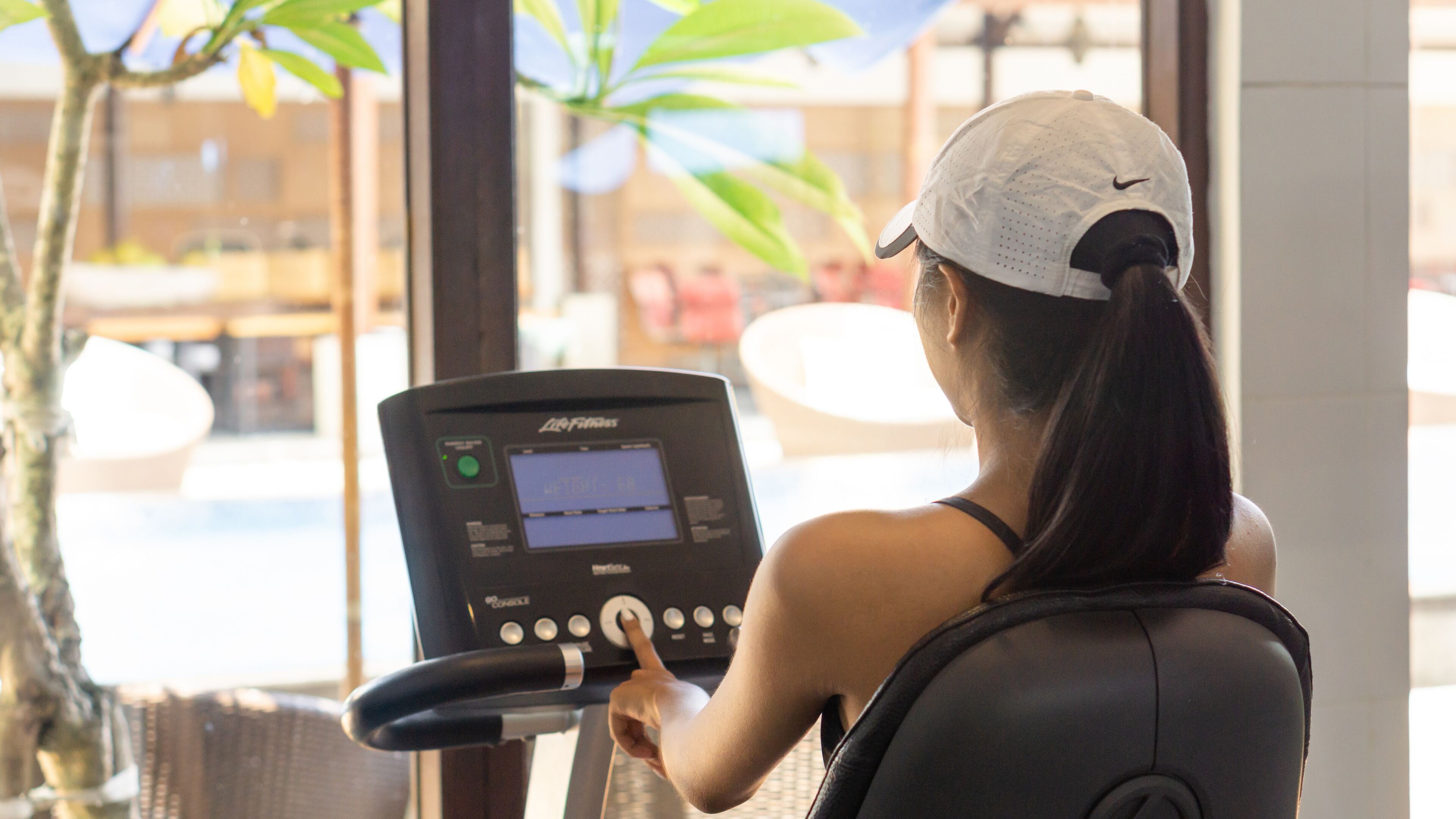 Una mujer usando una bicicleta estática con pantalla digital, en una habitación con ventanas amplias y plantas de interior.