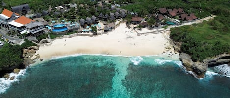 An aerial view of a beach resort with a sandy beach, clear turquoise waters, and several bungalow-style accommodations with thatched roofs.