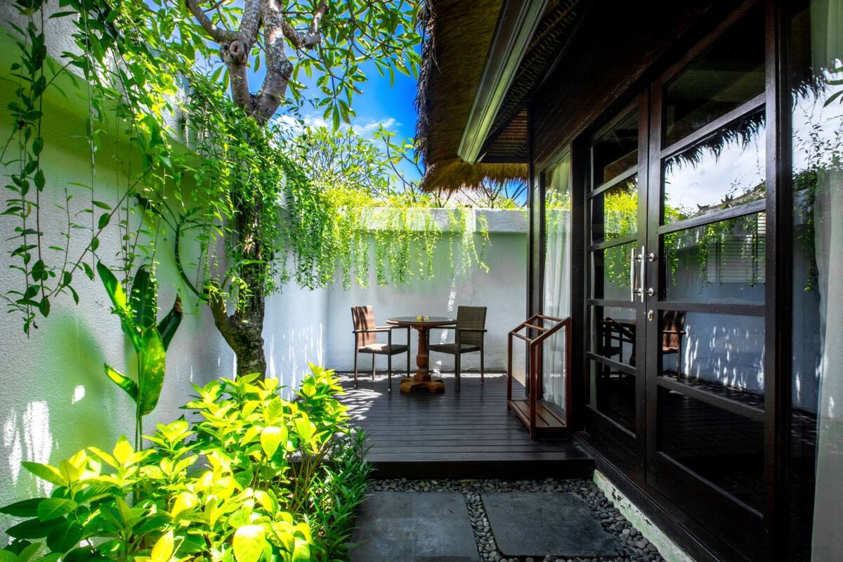 Une terrasse avec une table et des chaises, entourée de verdure et surmontée d’une structure à toit de chaume.
