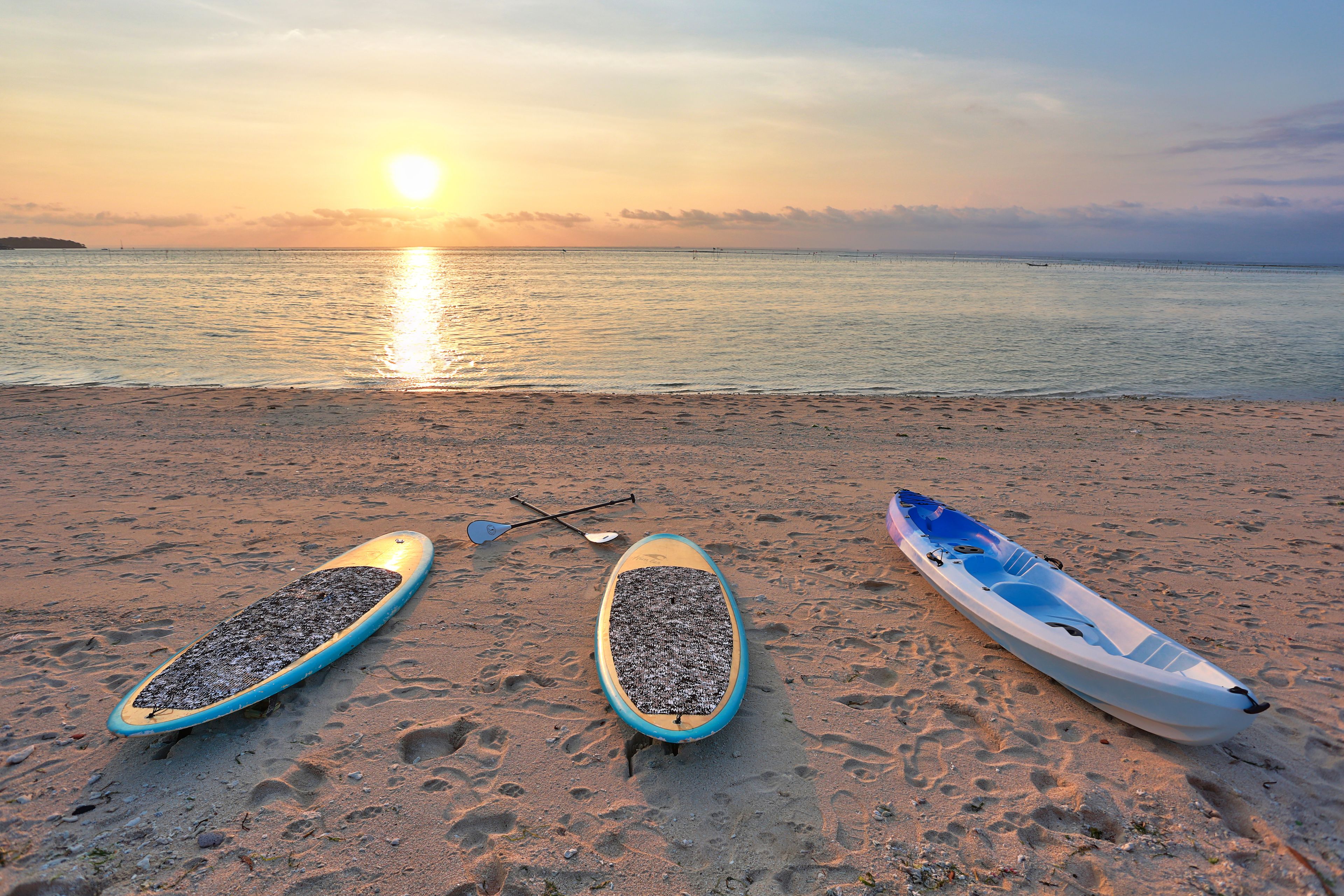 Una playa con dos tablas de remo y un kayak, y el sol se pone sobre el agua.