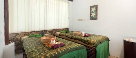A room with two beds, each with a green bedspread and a woven basket on the side. There is a stone wall and a window with blinds.