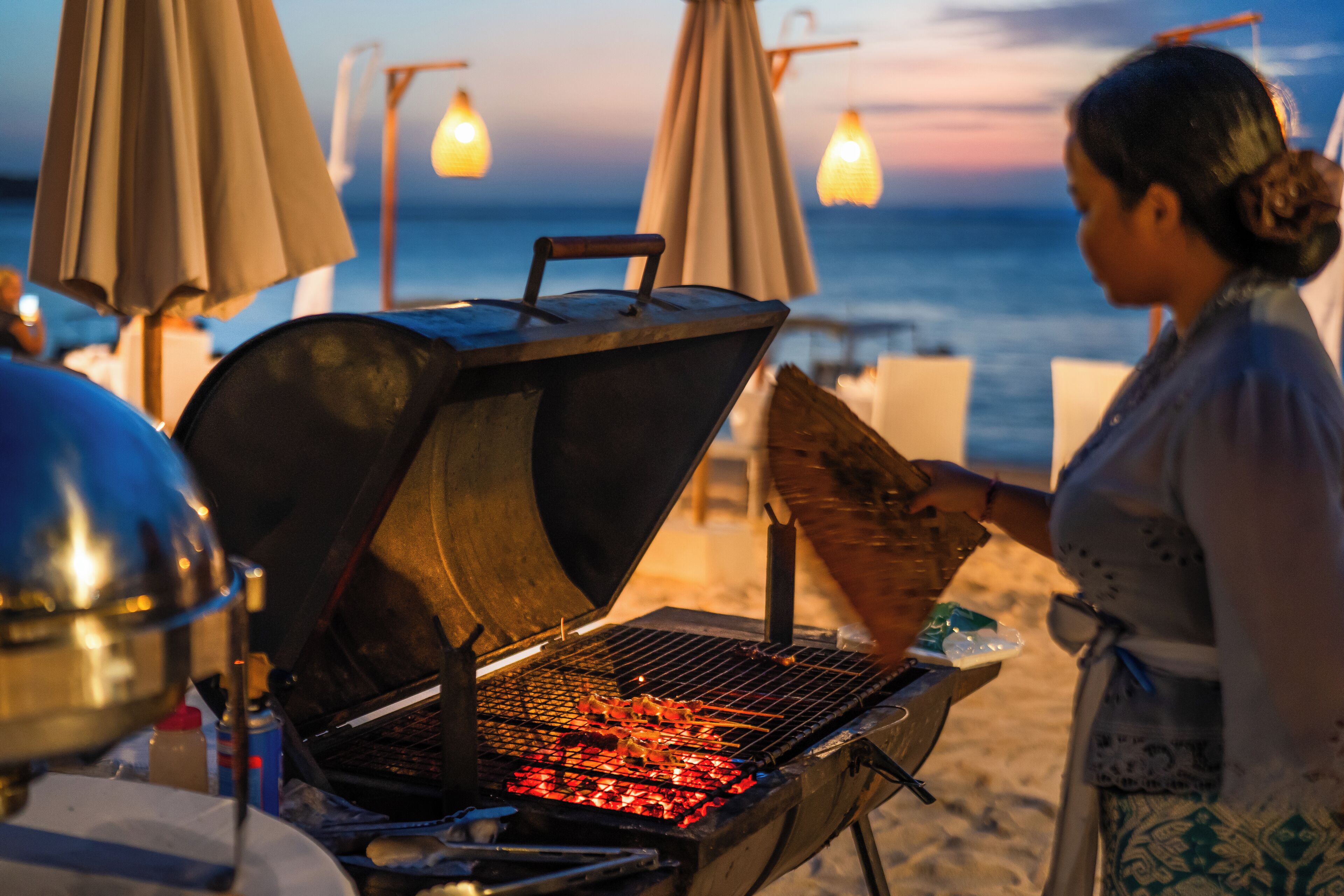 A barbecue grill with skewers on a beach with umbrellas and ocean in the background.