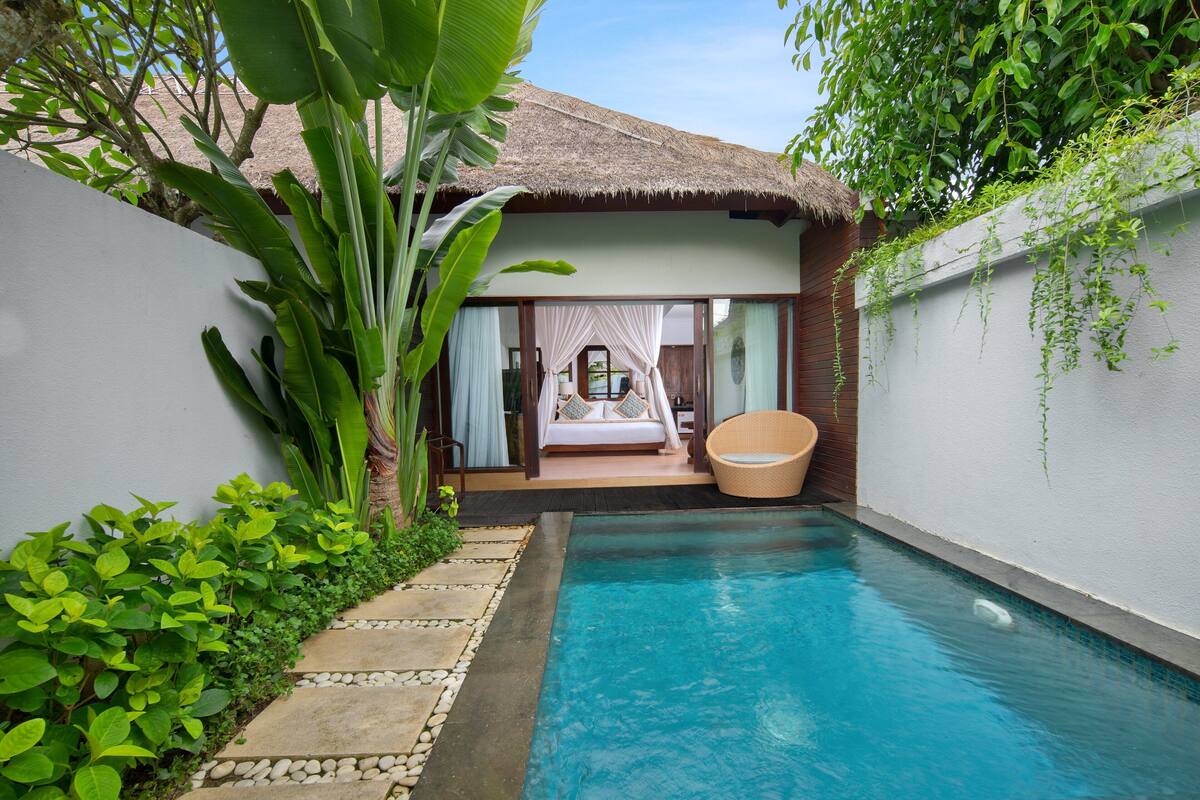 Un espace piscine privé comprenant un lit et une chaise, entouré d’une végétation luxuriante et abrité par une structure à toit de chaume.