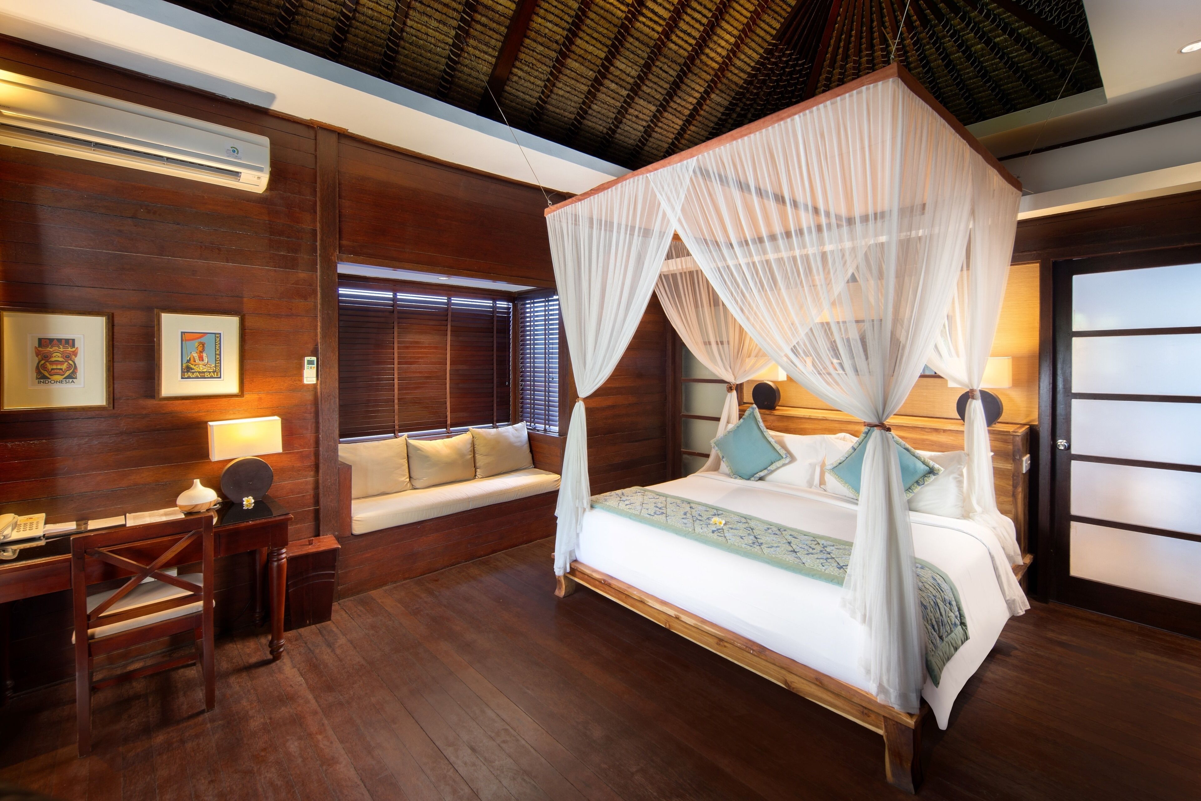 A spacious bedroom with a four-poster bed, wooden walls, a desk with a chair, and a window with blinds.