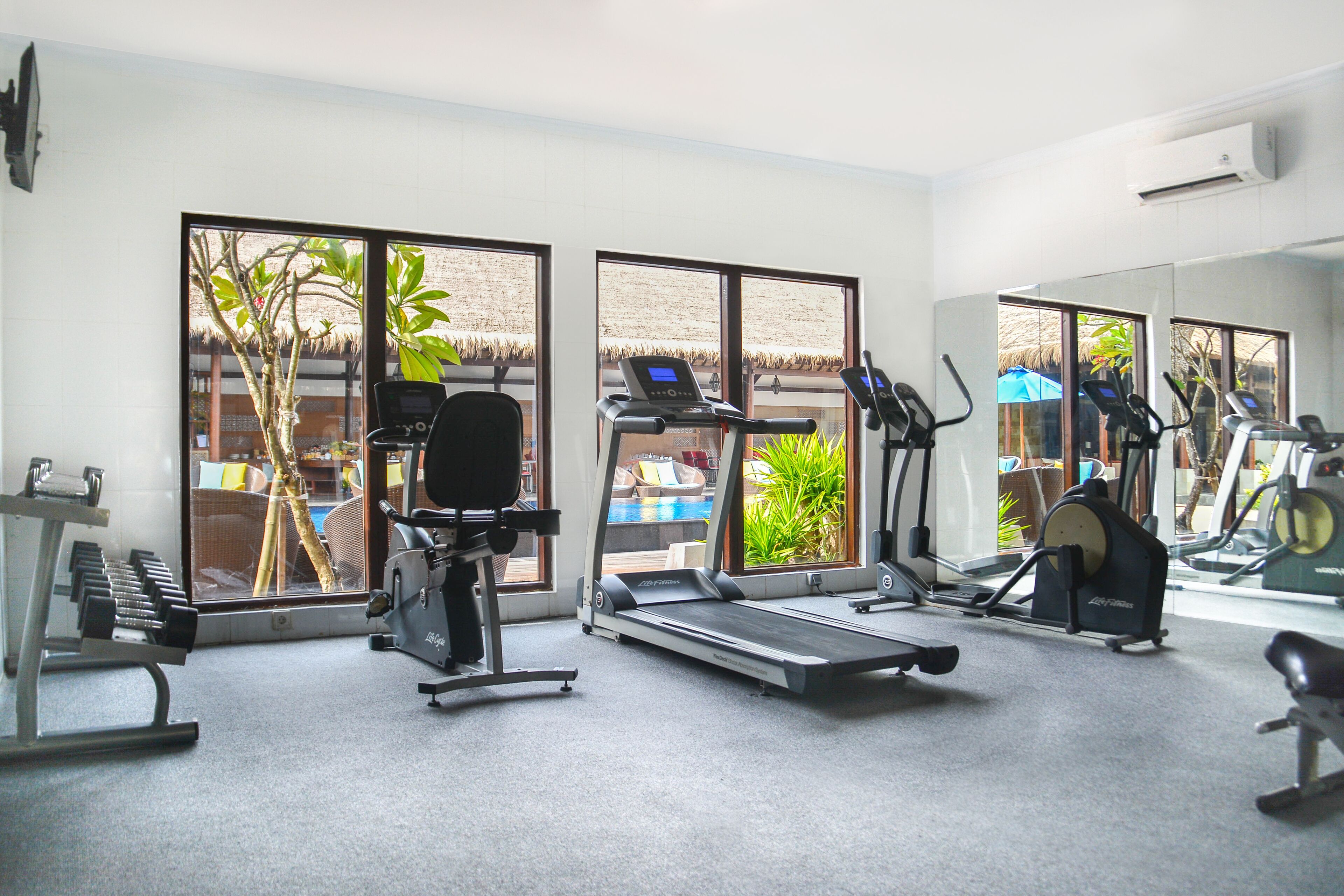 Een sportschool met cardioapparatuur, krachttrainingsmachines en grote ramen met uitzicht op een zwembad en palmbomen.