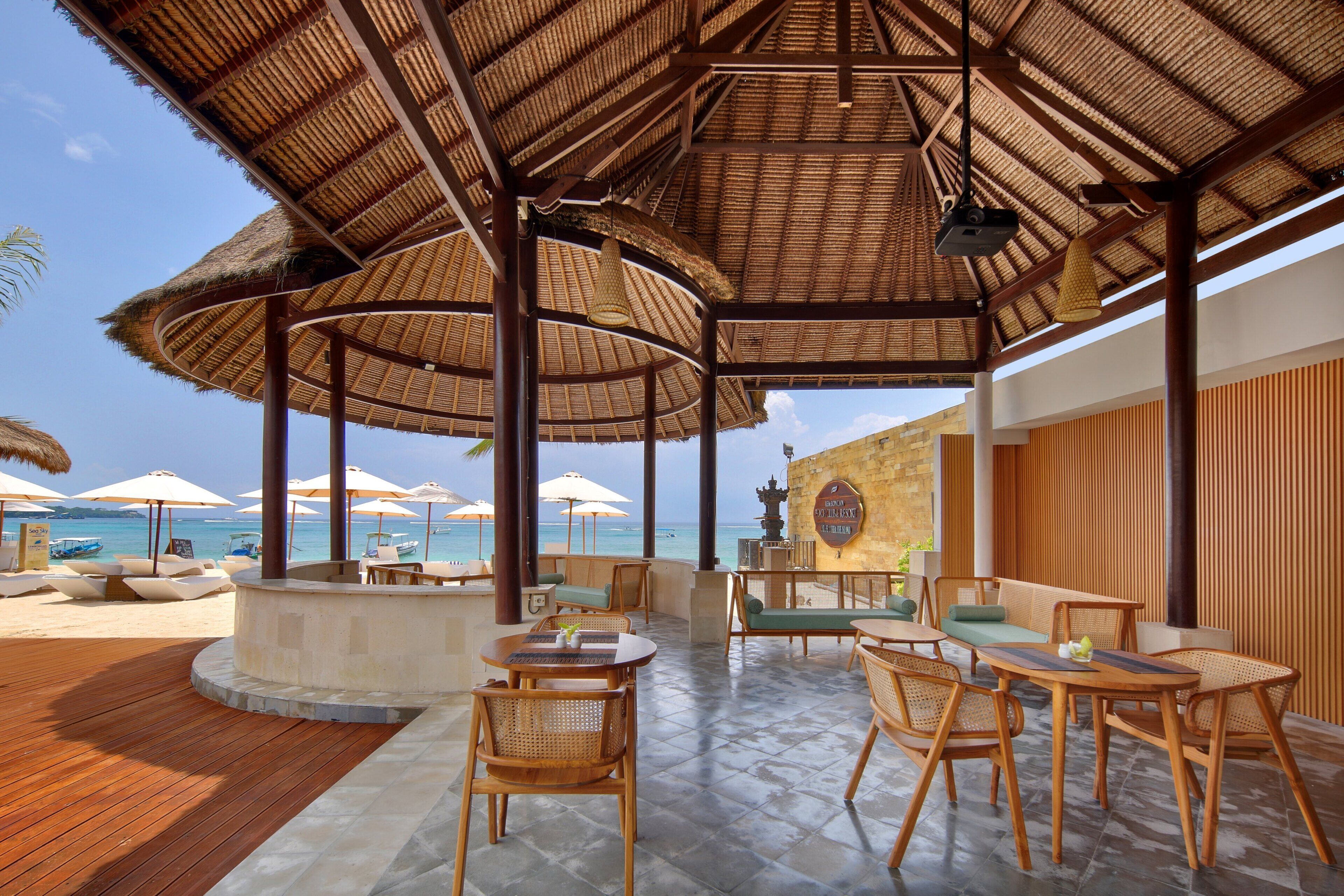 A covered outdoor seating area with wooden furniture and umbrellas, overlooking a beach.