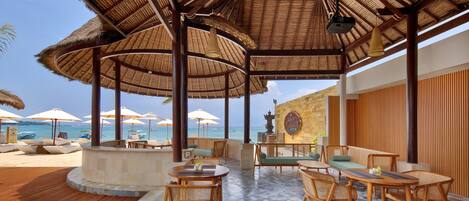 A covered outdoor seating area with wooden furniture and umbrellas, overlooking a beach.