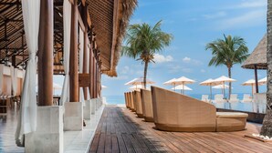 Um deck de madeira com móveis de vime e guarda-sóis, com vista para uma piscina e palmeiras.