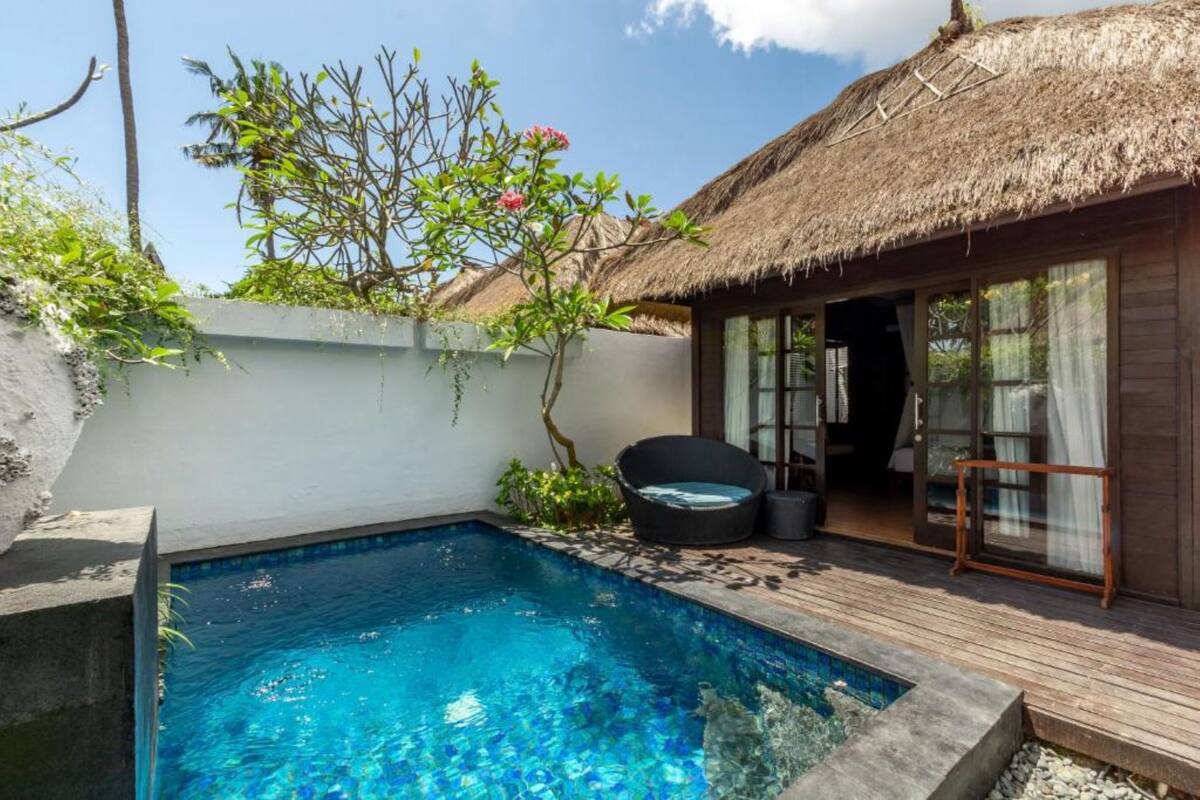 Une piscine avec une structure adjacente dotée d’un toit de chaume, une terrasse en bois et une porte vitrée donnant accès à une pièce.