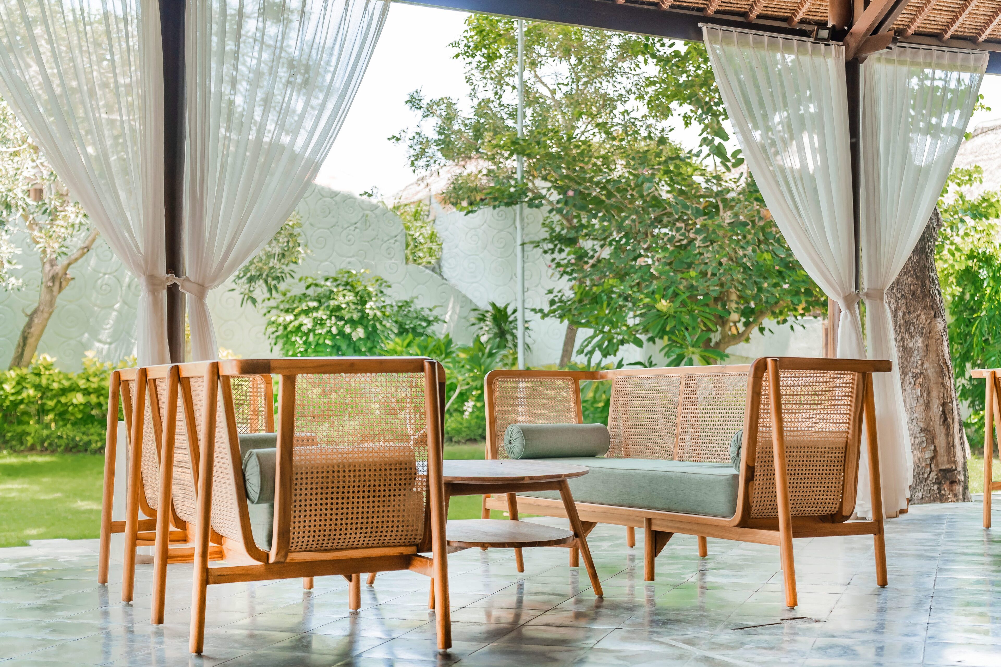 An outdoor seating area with wicker furniture and a small table, surrounded by a pergola with white curtains.