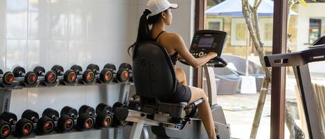 Una mujer hace ejercicio en bicicleta estática en un gimnasio, con una hilera de pesas y una cinta de correr al fondo.