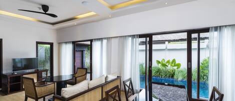 A modern living room with a dining area, large windows, a flat-screen TV, and a ceiling fan.