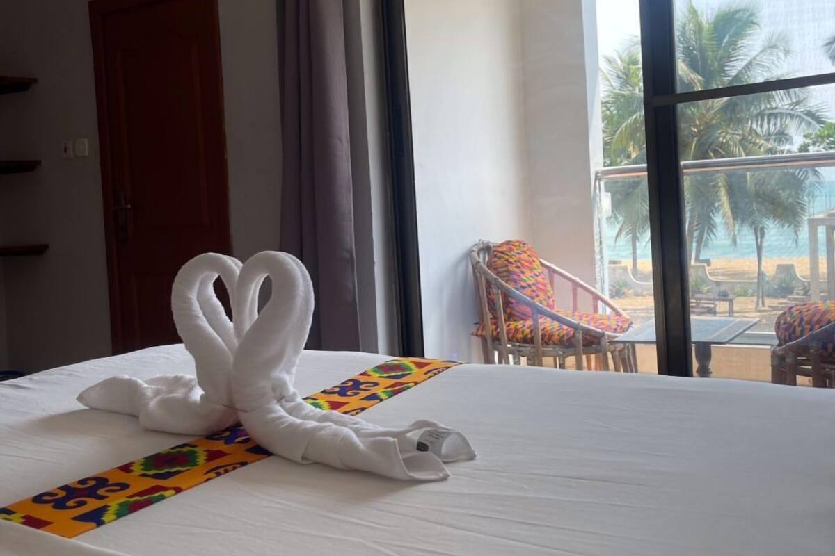 A hotel room with a bed, a large window with a view of palm trees and a beach, and a chair with colorful cushions.