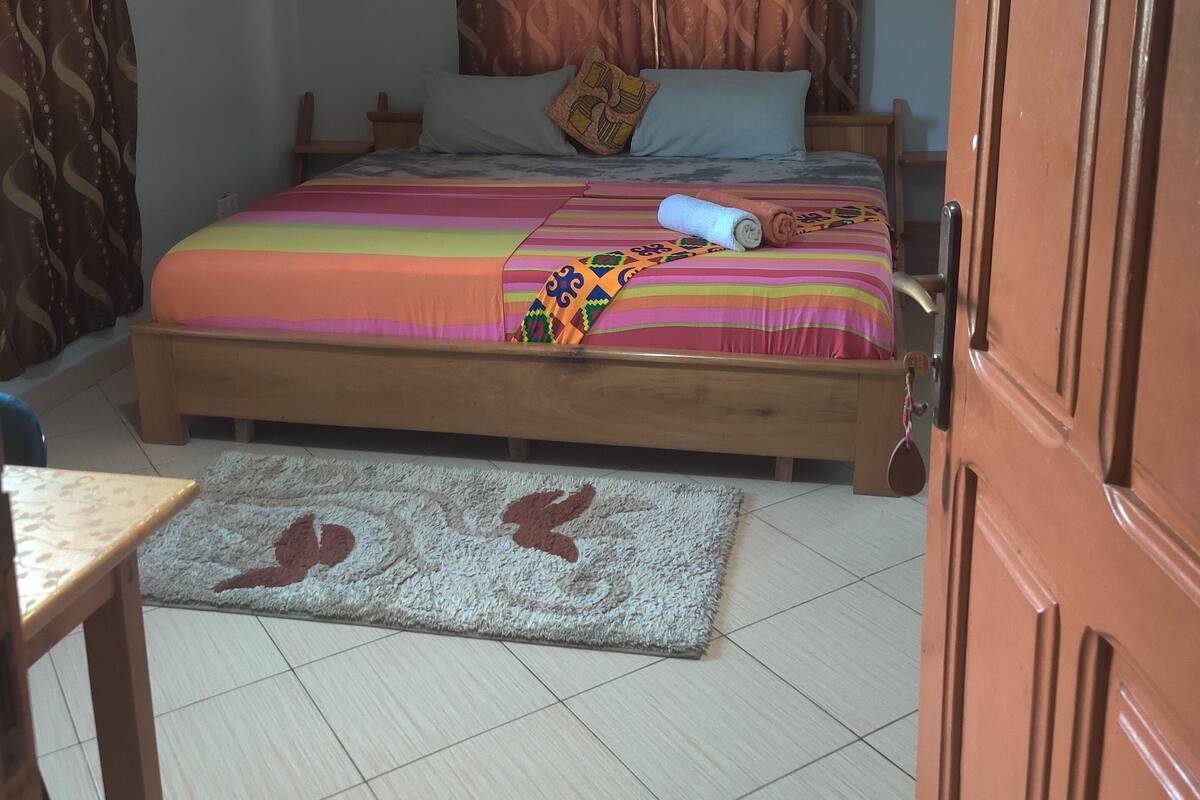 A bedroom with a bed, curtains, a ceiling fan, a wooden door, and a tiled floor.