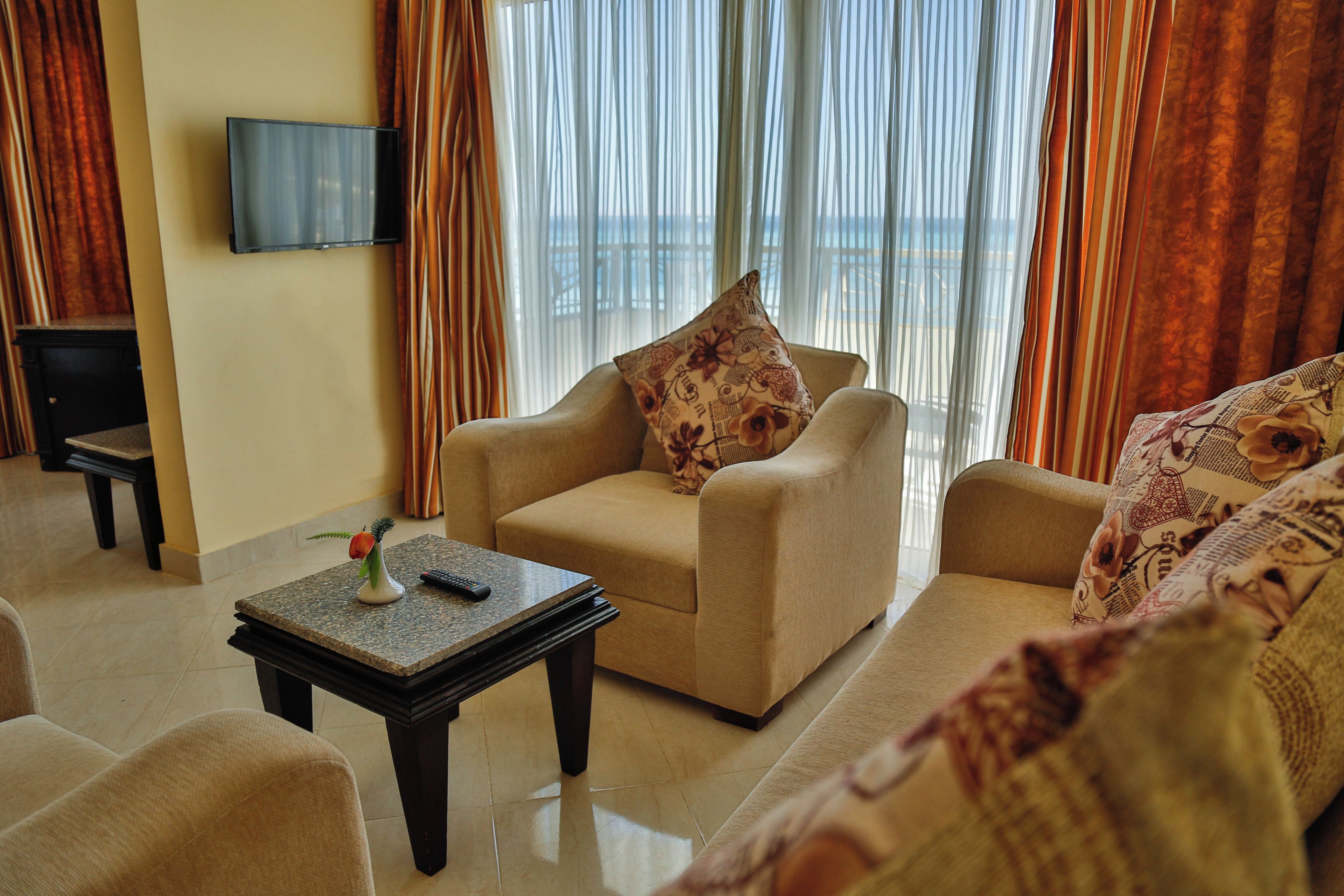A living room with a beige sofa, a small table with a vase and remote, and a TV on the wall.