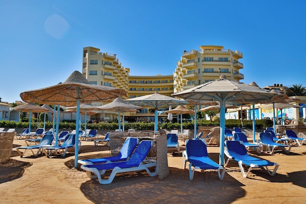 A beachside resort with sun loungers and umbrellas, a pool, and a hotel building in the background.