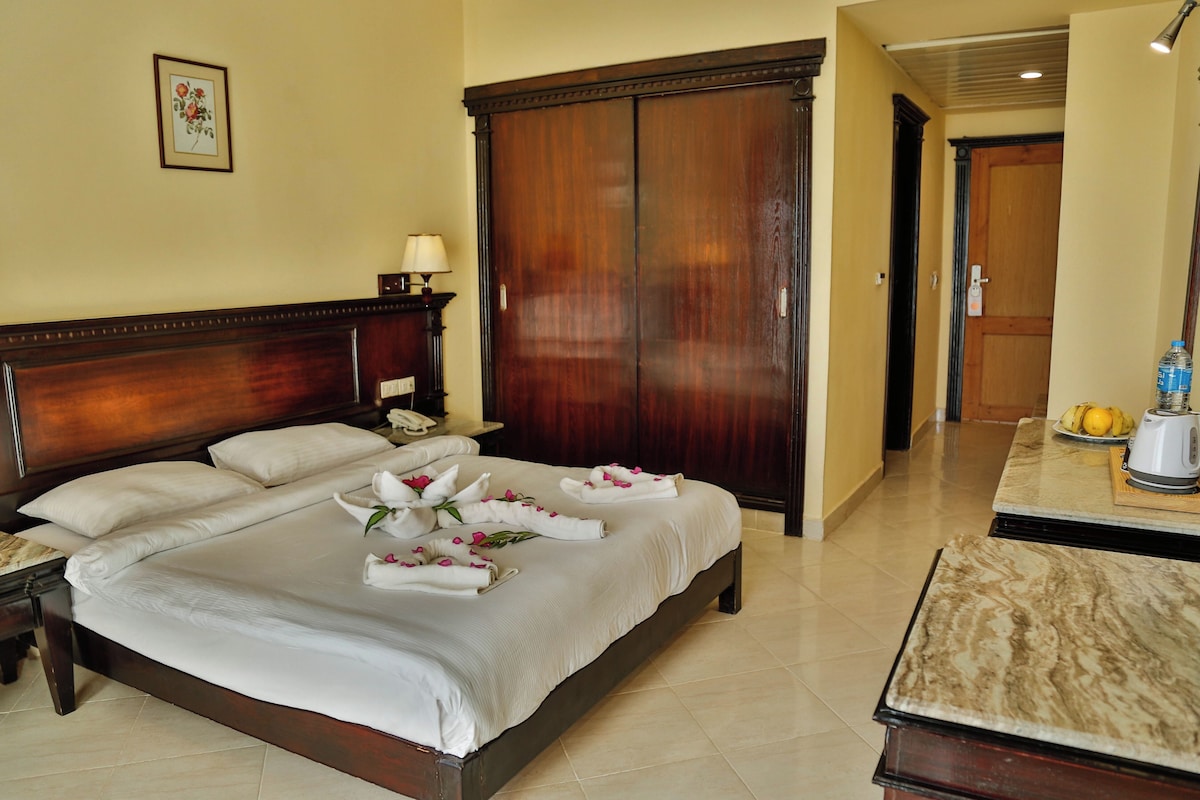 A hotel room with a large bed, a wooden wardrobe, a bedside table with a lamp, and a small table with a kettle and fruits.
