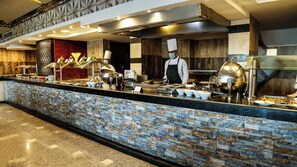 Montaje de bufé con diversos platos y cubiertos, un chef con uniforme y una cocina moderna.