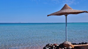 Klares, blaues Meer mit einer strohgedeckten Sonnenschirmkonstruktion an einer felsigen Küste.
