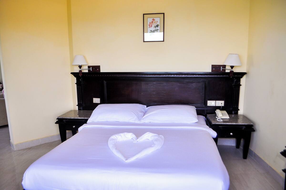 A hotel room with a large bed, two bedside tables, a heart-shaped towel on the bed, and a painting on the wall.