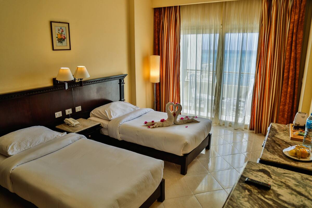 A hotel room with two beds, a bedside table with a lamp, a telephone, and a window with curtains.