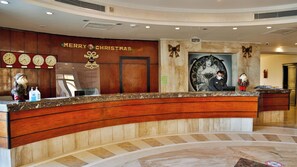 Un mostrador de recepción con detalles de madera, un cartel que dice 'Feliz Navidad', relojes mostrando diferentes ciudades y una recepcionista.