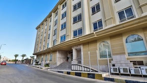 A multi-story hotel with a central entrance, a staircase leading to the entrance, and a row of air conditioning units on the ground floor.