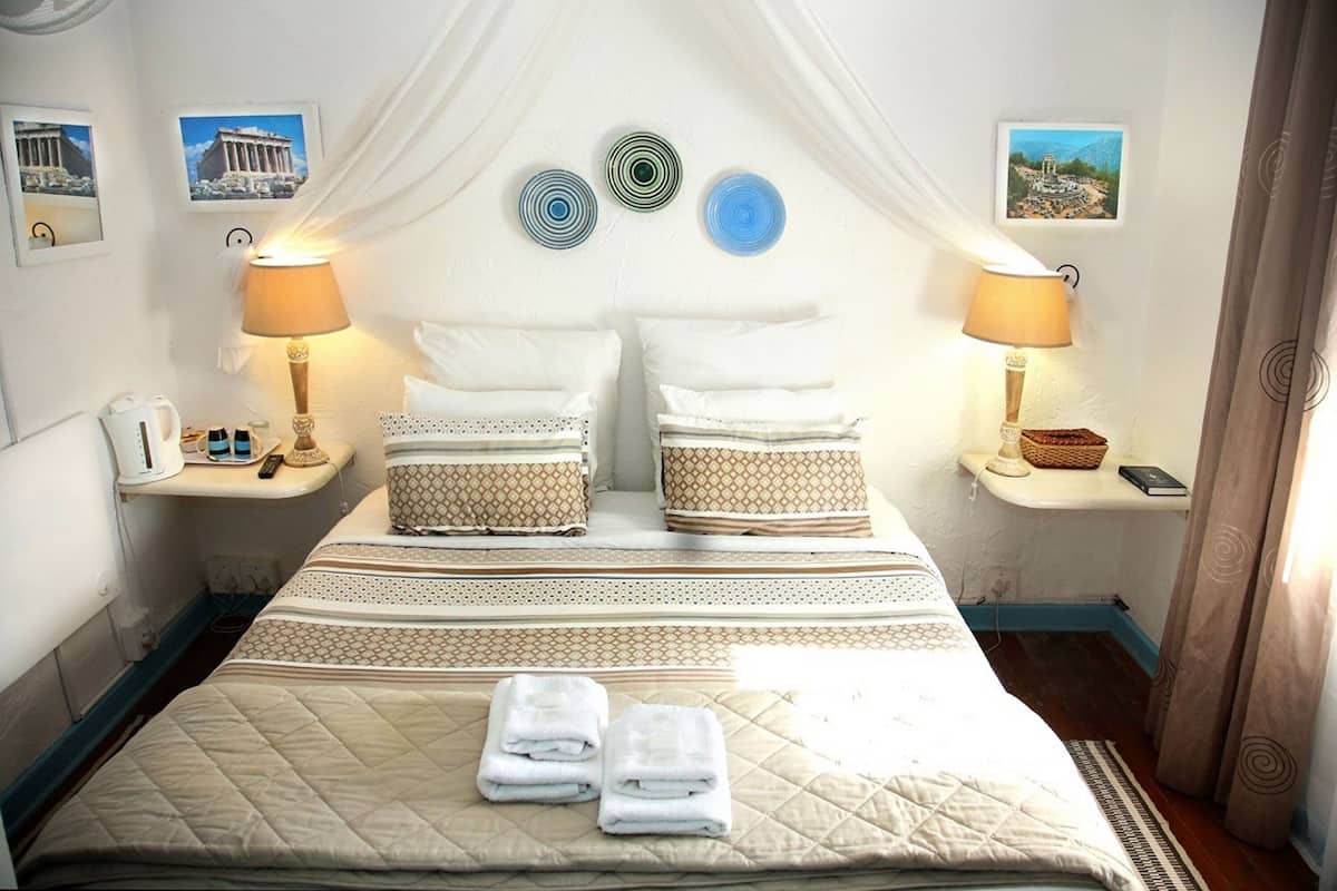A neatly arranged hotel room with a bed, two bedside tables, a bedside lamp, a wall-mounted phone, and a small shelf with a book and a basket.