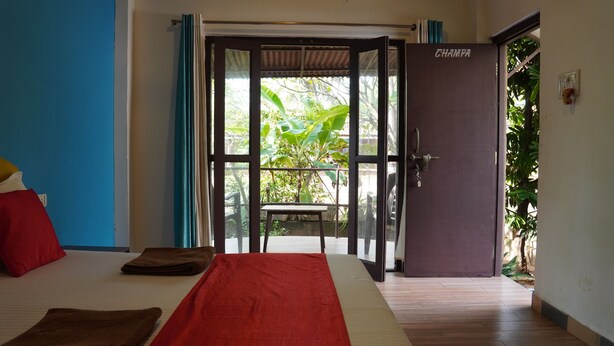 Une pièce avec un lit, une couverture rouge et beige, un plancher en bois, une porte portant l’inscription « CHAMPA » et une vue sur la verdure à l’extérieur.