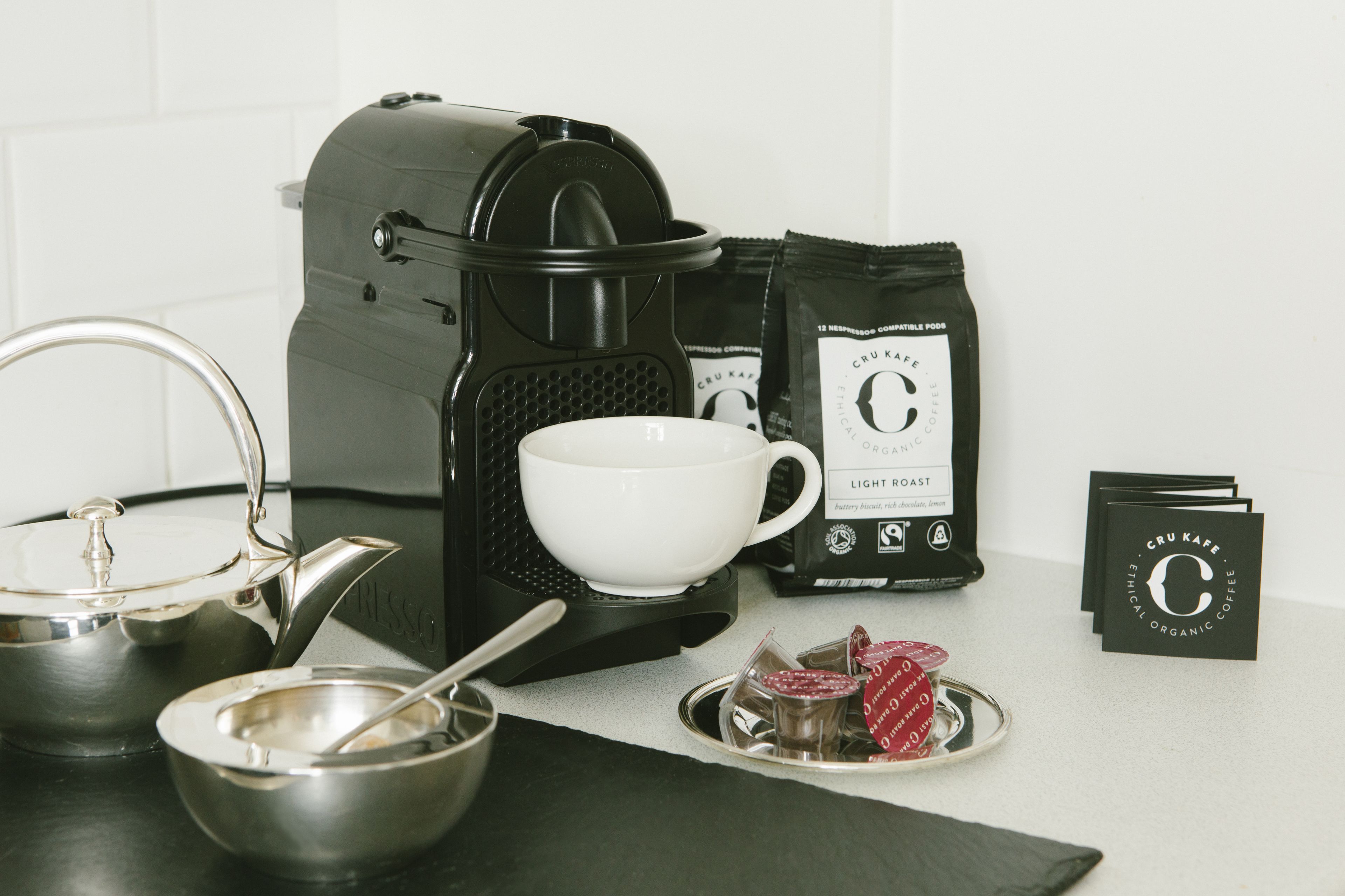 A coffee-making setup with a coffee maker, a white cup, a kettle, a tea pot, a spoon, a tray with tea bags, and a bag of coffee.