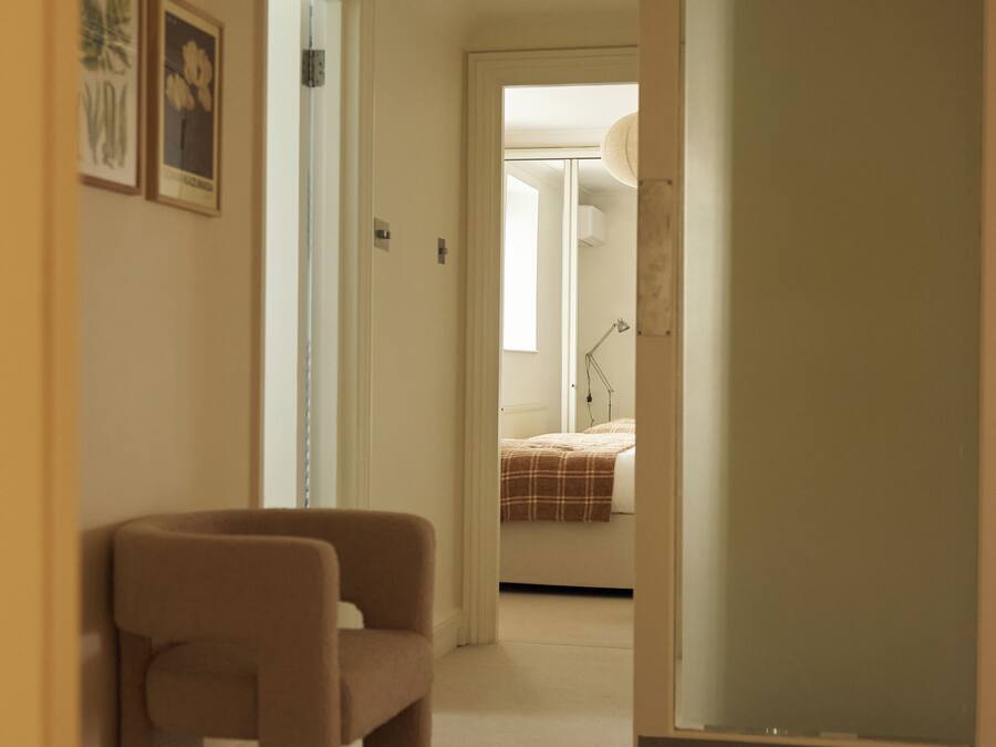Un couloir avec un tapis beige, une chaise marron et un lit à l’arrière-plan.