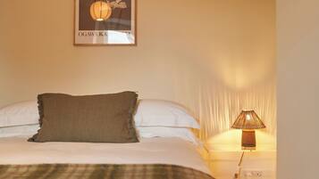 A neatly made bed with a brown throw pillow, a white pillow, and a plaid blanket. There is a framed picture of flowers on the wall above the bed. A lamp with a striped lampshade is on the nightstand beside the bed.