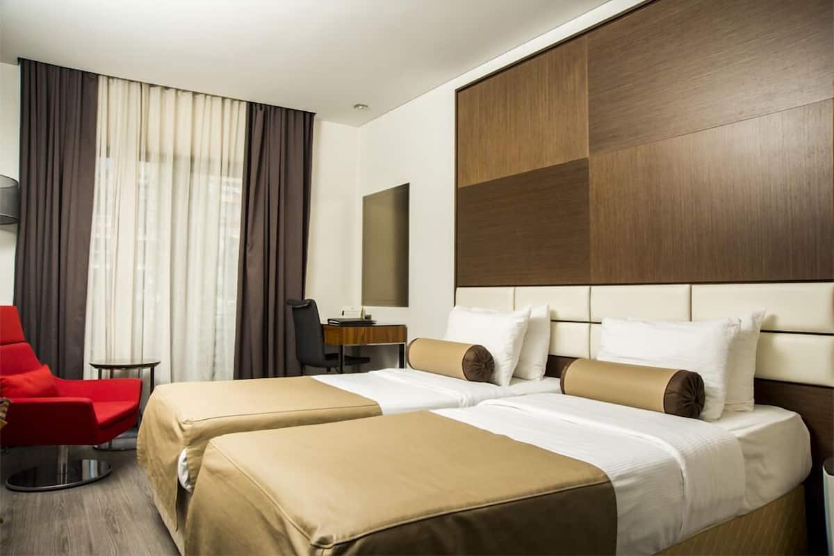 Une chambre d’hôtel avec deux lits, un fauteuil rouge, un bureau et une grande fenêtre avec des rideaux.