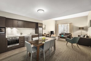 A modern kitchen with dark wood cabinets, a dining area with a wooden table and chairs, a living room with a sofa and a TV, and a large window with curtains. - Quest Albury On Townsend (Albury)
