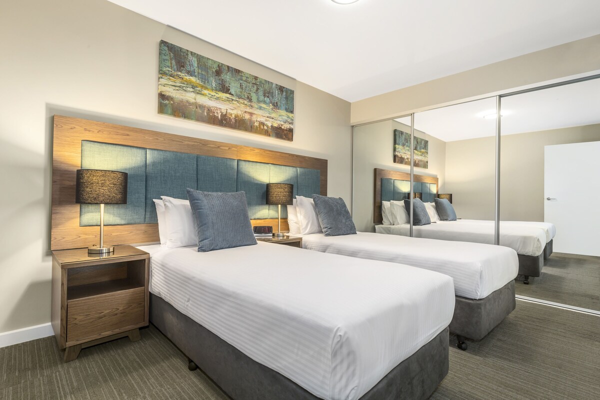 A hotel room with two beds, a wooden headboard, a nightstand, a lamp, and a painting on the wall.