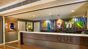 A hotel reception area with a wooden reception desk labeled "QUEST" and various colorful artworks on the walls. - Quest Albury On Townsend (Albury)