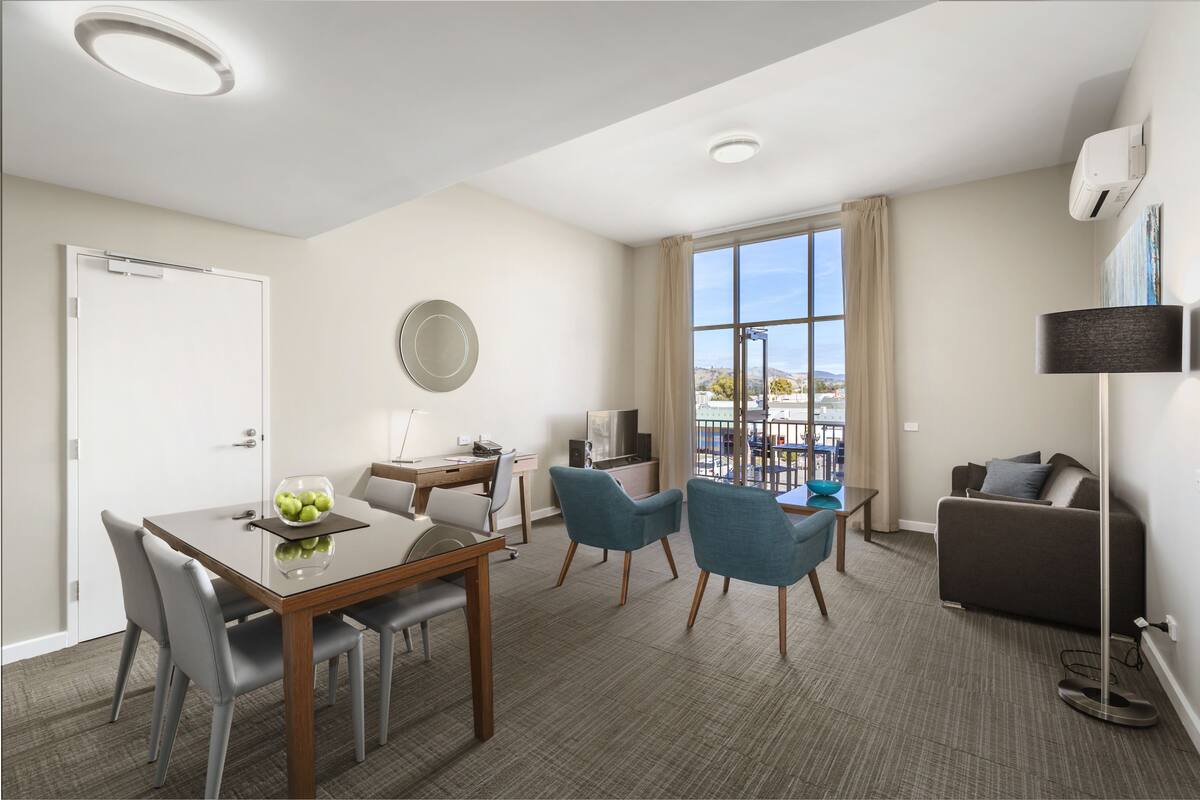 Quarto de hotel moderno com área de refeições, sofá, TV e uma grande janela com vista.