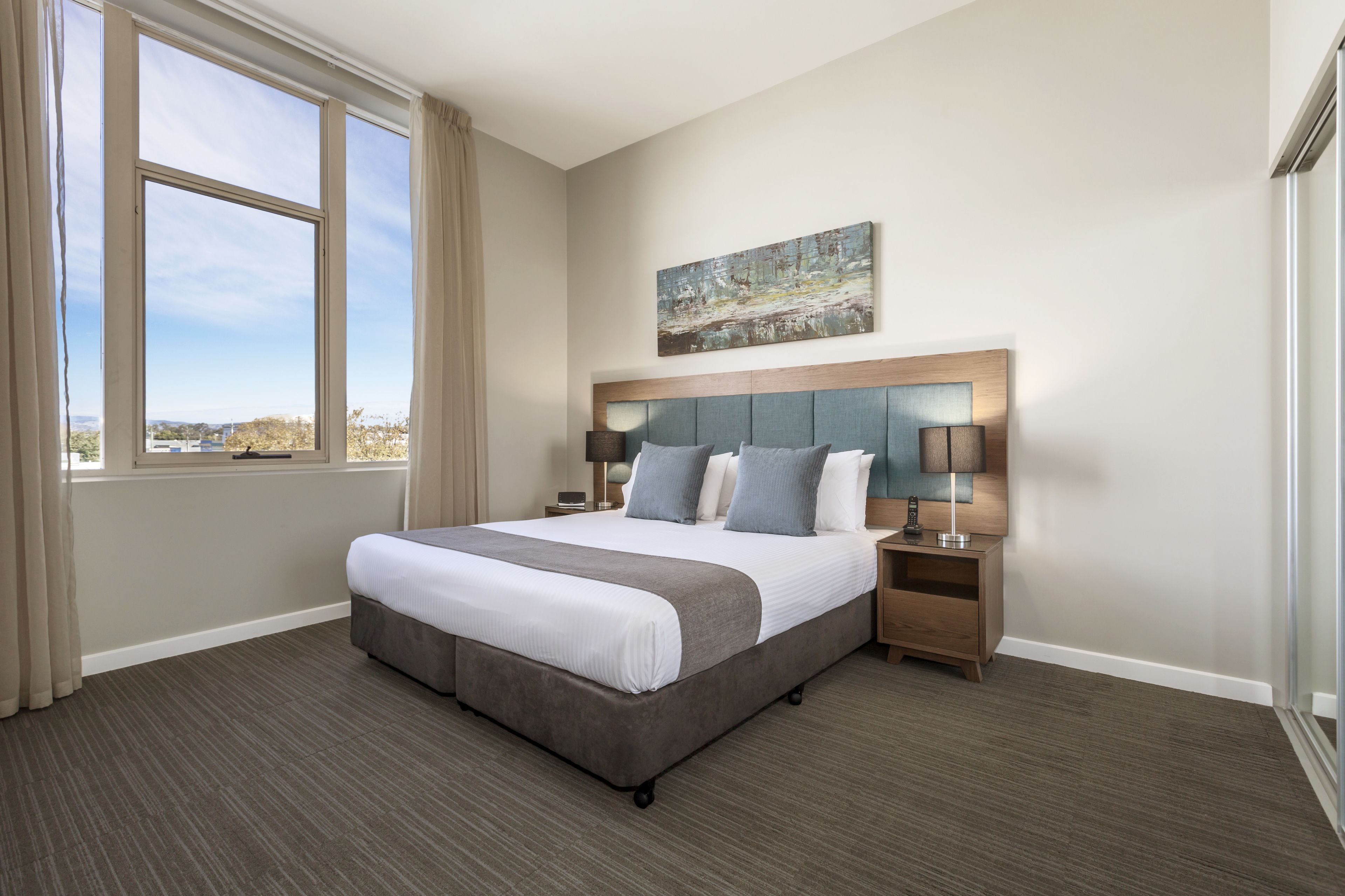 A hotel room with a large bed, a headboard with a painting, two bedside tables with lamps, and a view of the city through large windows.