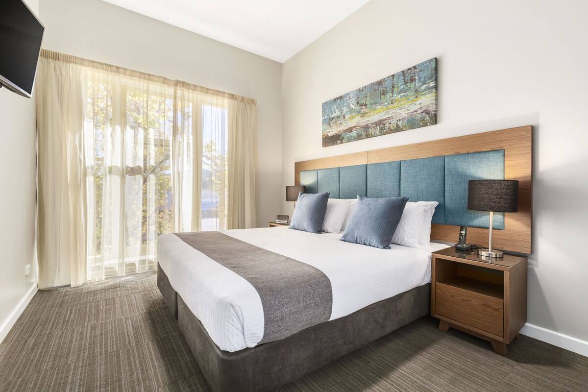 Quarto de hotel com uma cama grande, cabeceira com painel azul, mesa de cabeceira com abajur e um quadro na parede.