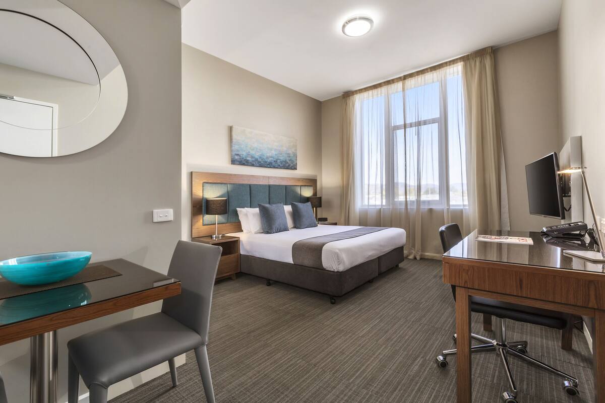 Quarto de hotel com cama, escrivaninha, cadeira, televisão e uma janela grande com cortinas.