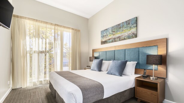A hotel room with a large bed, a nightstand with a lamp, a painting on the wall, and sheer curtains on the window. - Quest Albury On Townsend (Albury)