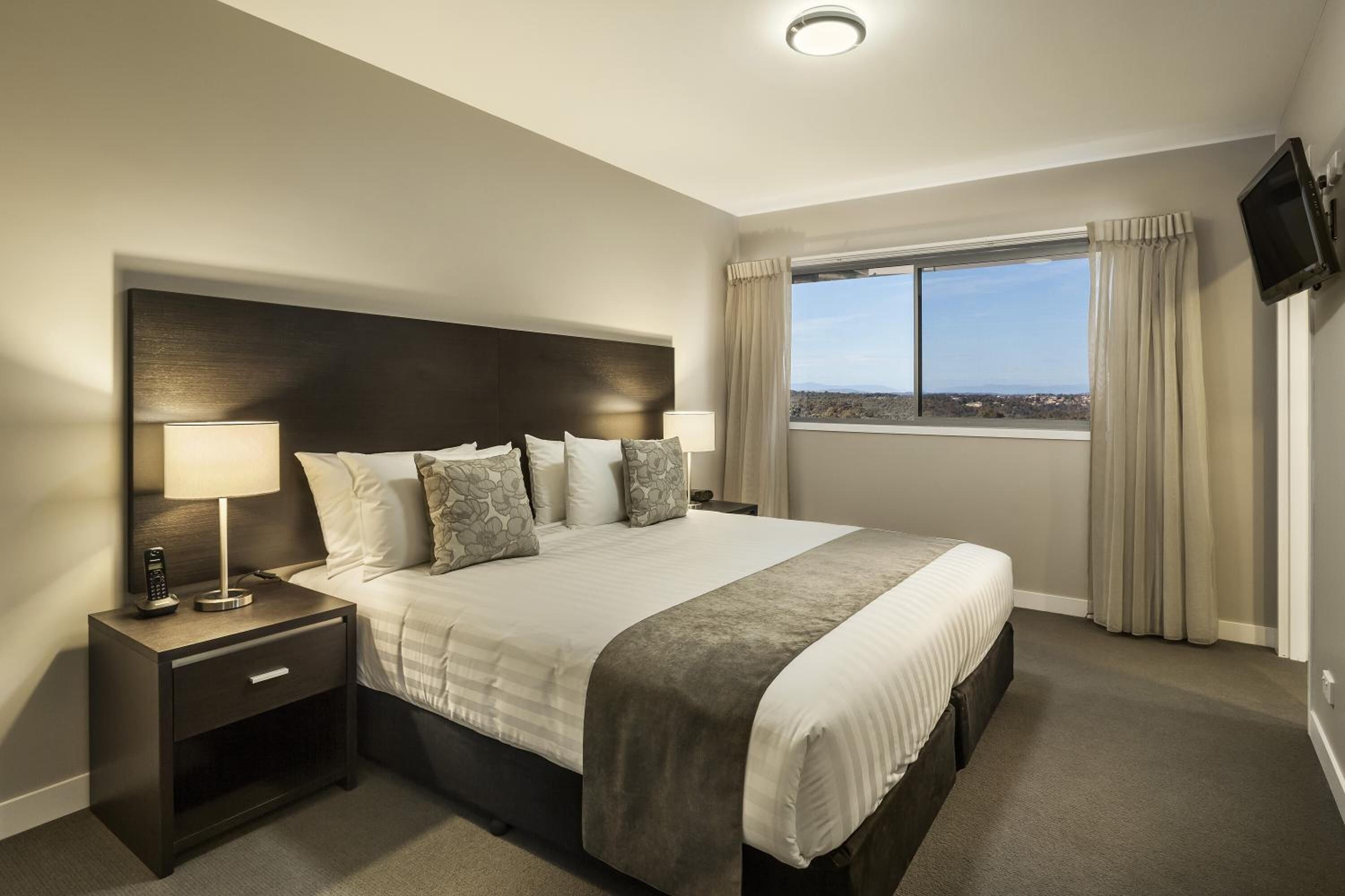 Ein Hotelzimmer mit einem großen Bett, zwei Nachttischen mit Lampen, einem an der Wand montierten Fernseher und einem Fenster mit Vorhängen.